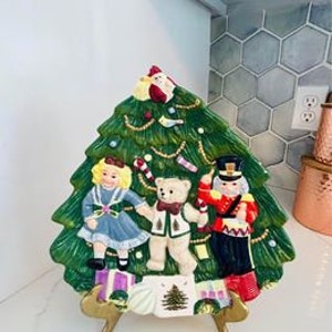 May include: A ceramic Christmas tree-shaped serving plate with a green tree, a girl in a blue dress, a teddy bear, and a nutcracker. The plate has a gold stand.