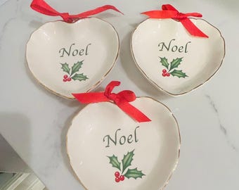 May include: Three white heart-shaped ceramic dishes with gold trim. Each dish has a red ribbon and a green holly sprig with red berries. The word "Noel" is written on each dish.