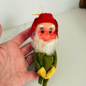 May include: A small elf ornament featuring a red felt hat, a white synthetic beard, and a green felt body with yellow cuffs. The figure has a painted face with blue eyes, arched white eyebrows, and a gold hanging loop at the top.