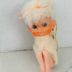 May include: A small hanging ornament featuring a doll with white fuzzy hair, blue eyes, and rosy cheeks. The doll wears a white fabric garment and has pink legs. A thin orange string is attached to the top of the head for hanging purposes.