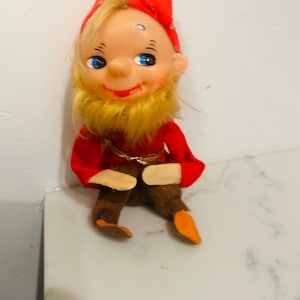 May include: A vintage gnome doll with a red hat and outfit, a yellow beard, and blue eyes. The doll has a smiling expression and is sitting down. The doll's shoes are orange and brown.