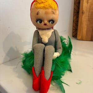 May include: A doll with a large round head, yellow yarn hair, and a red cap. It has painted blue eyes with long lashes and red lips. The doll wears a gray felt outfit with a white collar and red pointed shoes, sitting atop a cluster of green feathers.