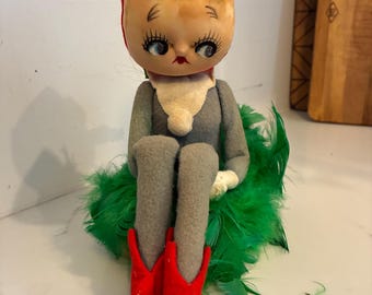 May include: A doll with a large round head, yellow yarn hair, and a red cap. It has painted blue eyes with long lashes and red lips. The doll wears a gray felt outfit with a white collar and red pointed shoes, sitting atop a cluster of green feathers.