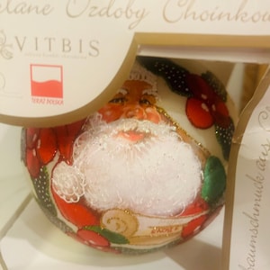 May include: A round Christmas ornament featuring Santa Claus with a white beard and red suit. The ornament is decorated with red and green accents and is packaged in a box.