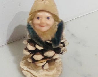 May include: A whimsical figurine of a gnome crafted from a pine cone. The gnome has a smiling face, a tan hat, and a green collar. The base is a light brown color.