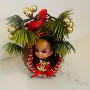 May include: A vintage Christmas ornament featuring a doll-like figure with blonde hair, dressed in red and green, holding a golden harp. The ornament is surrounded by green palm fronds, gold berries, and a red ribbon.