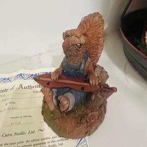 May include: A Cairn Studio figurine of a squirrel playing a stringed instrument. The squirrel wears blue overalls and sits on a tree stump base. The figurine has brown and tan coloring. A certificate of authenticity is partially visible.