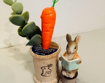 May include: A whimsical Easter-themed display featuring a bright orange carrot with green leaves, a faux succulent, and a figurine of a rabbit reading a book. The carrot is placed in a vintage thread spool.