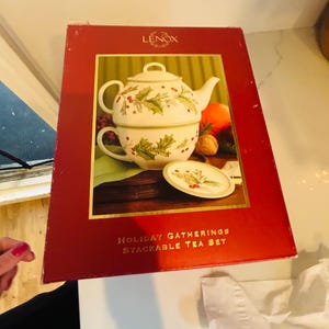 May include: A red box featuring a Lenox Holiday Gatherings stackable tea set. The tea set includes a teapot, teacup, and saucer, all decorated with holly and berry designs. The box has the words "HOLIDAY GATHERINGS STACKABLE TEA SET" printed on it.