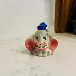 May include: A small ceramic elephant figurine featuring large pink ears, a blue hat, and a white collar with green and red trim. The elephant has a light gray body, painted black eyes with long eyelashes, and rosy pink cheeks on a white face.
