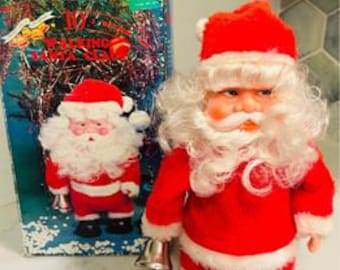 May include: A vintage, battery-operated, walking Santa Claus toy. He is wearing a red suit with white trim, a white beard, and a red hat. He is holding a bell in his left hand and has a smiling face.