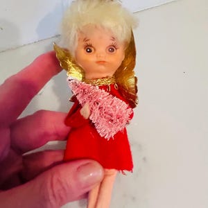 May include: Vintage Christmas angel ornament. The doll has a plastic face, blonde hair, and gold-colored wings. She wears a red dress and holds a pink, fuzzy tree. A gold halo sits atop her head. The ornament is held by a hand.
