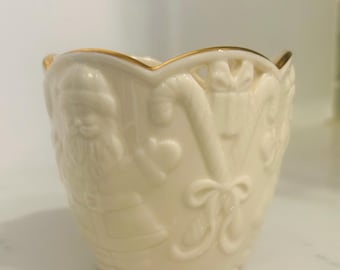 May include: A white ceramic candle holder with a gold rim. The holder is decorated with a Christmas scene featuring Santa Claus, candy canes, and a bow.