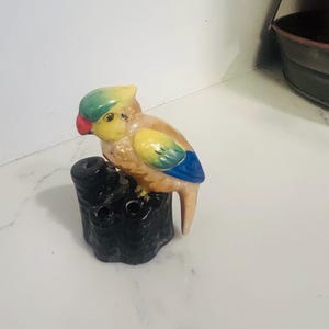 May include: A ceramic figurine of a parrot perched on a black, cylindrical base with multiple circular openings. The bird features a green crest, a red beak, a yellow face, and a body with yellow, green, and blue wing details on a tan background.