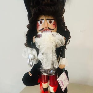 May include: A wooden Captain Hook nutcracker figurine. The figurine has a black hat with white feathers, a curly brown wig, and a white lace collar. It wears a black coat, red pants, and red shoes with bows. The base reads "Captain Hook".