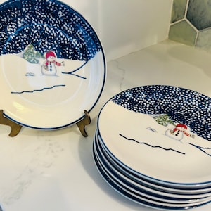 May include: Set of six white ceramic plates with a blue and white snowflake design. Each plate features a snowman illustration.