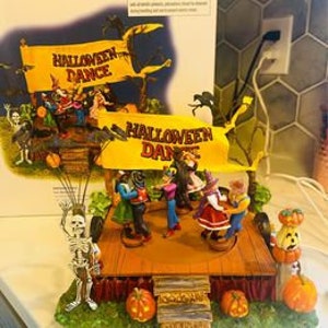 Op de afbeelding: Een Halloween-dorpsdecor met een podium waarop een groep skeletten danst. Het podium is versierd met pompoenen en een banner met de tekst "Halloween Dance".