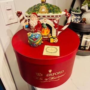 May include: A ceramic teapot shaped like a fireplace with a Santa Claus figure holding a sack of gifts, a lit hearth, and candy cane handles. It sits on a round red Waterford Holiday Heirlooms box with gold text and includes a small square product card.