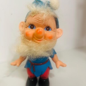 May include: A vintage gnome figurine with a blue hat and tunic, red pants, and black boots. The gnome has a white beard, rosy cheeks, and a large orange nose. A string is attached to the top of the hat.