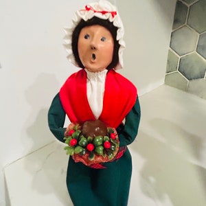 May include: A Christmas carol figurine of a woman wearing a white bonnet, red and green dress, and holding a bowl of holly berries.