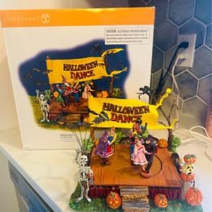 Op de afbeelding: Een muzikaal dorpssc&egrave;ne met Halloween-thema met een draaiende dansvloer, skeletten, pompoenen en een banner met de tekst "Halloween Dance".