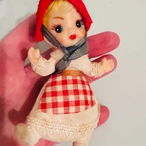 May include: A vintage doll ornament with a red bonnet, gray scarf, and a white dress with a red and white checkered skirt. The doll has blonde hair, rosy cheeks, and is holding its arms out. The ornament is likely made of fabric and plastic.
