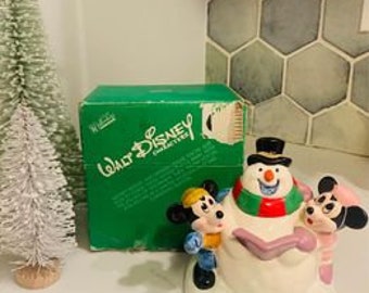 May include: A vintage ceramic figurine of Mickey Mouse and Minnie Mouse building a snowman. The figurine is painted in bright colors and features a green, red, and white scarf on the snowman. The figurine is in its original box, which is green and features the Walt Disney logo.