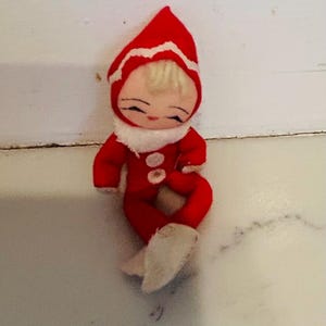 May include: A vintage doll dressed in a red jumpsuit and matching pointed hat with white trim. The doll has a white collar, white boots, and a serene expression. The doll is a collectible item.