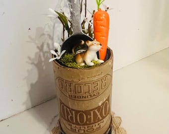 May include: A decorative arrangement featuring a cylindrical container with the text "OXFORD" and "RECORD". Inside, a small dog figurine sits next to a carrot and faux greenery. The arrangement is on a lace doily.