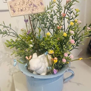 May include: A blue enamelware cup filled with greenery, colorful flowers, and a white ceramic bunny. A small sign that reads "Happy Easter" is placed in the arrangement. The cup has a handle and sits on a white surface.