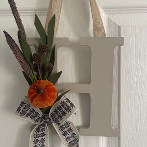 Puede incluir: Una letra H de madera gris claro con un colgador de cinta de arpillera. La letra está decorada con una pequeña calabaza de terciopelo naranja, un lazo de cinta blanca con un estampado floral azul marino y una rama de vegetación artificial.