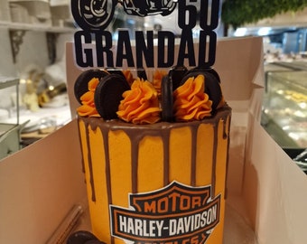May include: A Harley-Davidson themed cake with orange frosting and chocolate drips. The cake is decorated with a Harley-Davidson logo and a topper that says "60 GRANDAD" with a motorcycle silhouette. The cake is in a white box.