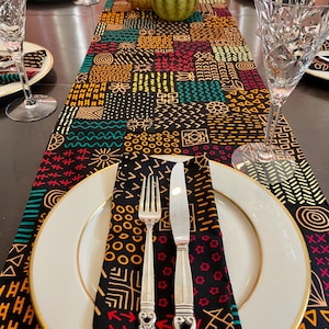 Może przedstawiać: Czarny bieżnik stołowy z kolorowym geometrycznym wzorem. Bieżnik jest wykonany z tkaniny i ma różne kształty i wzory. Bieżnik leży na drewnianym stole z białym talerzem, srebrnym widelcem i nożem oraz serwetką.