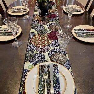Pode incluir: Uma mesa de jantar montada com um caminho de mesa estampado com um design floral em vermelho, verde e azul. Cada lugar inclui um prato branco com borda dourada, talheres e uma taça de vinho de cristal. Um vaso decorativo com vegetação está no centro.