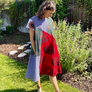 May include: A woman wearing a patchwork dress with a red, green, blue, and white pattern. The dress has a short sleeve and a loose fit. The woman is standing in a garden with green grass and a wooden fence in the background.
