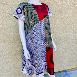 May include: A patchwork dress with a geometric pattern in shades of blue, green, red, and white. The dress has a round neckline and short sleeves. The text "IMAN" is visible on the dress.