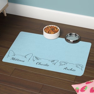 May include: A light blue pet food mat with three cat silhouettes and the names Mittens, Charlie, and Archie. A white ceramic bowl with pet food and a silver metal bowl are on the mat.