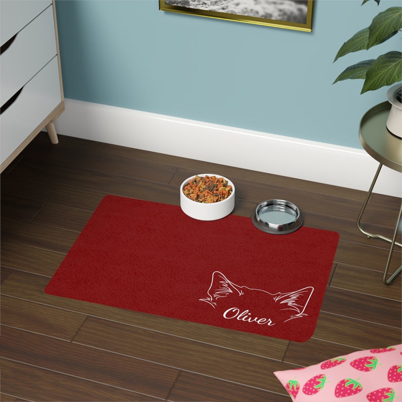May include: Red pet food mat with a white outline of a cat's face and the name "Oliver" printed on it. A white ceramic bowl with pet food and a silver metal bowl are on the mat.