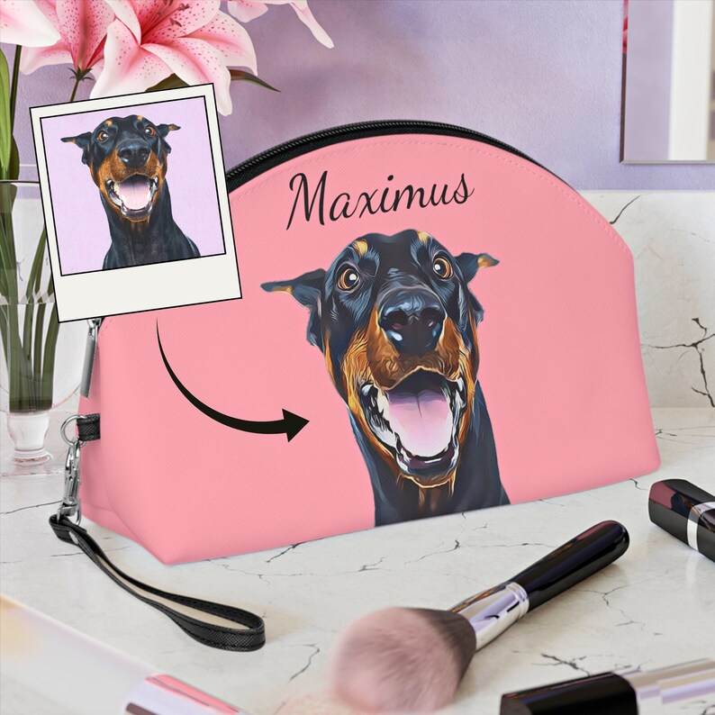 May include: Pink cosmetic bag featuring a Doberman Pinscher illustration with the name "Maximus" in black script. A smaller image of the dog is on a polaroid-style frame. The bag has a black zipper and wrist strap.