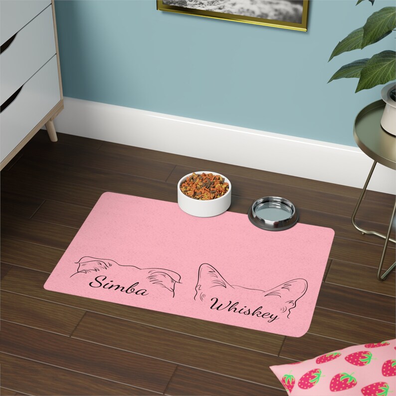 May include: Pink dog food mat with two dog silhouettes and the names Simba and Whiskey printed on it. A white ceramic bowl with dog food and a silver metal water bowl are on the mat.