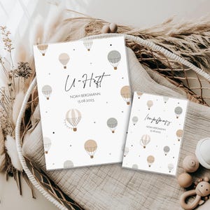 Könnte beinhalten: Zwei weiße Baby-Gesundheitshefte mit Illustrationen von Heißluftballons in Beige, Grau und Hellbraun. Das größere Buch trägt die Aufschrift "U-Heft" mit Namen und Datum. Das kleinere Buch trägt die Aufschrift "Impfpas".