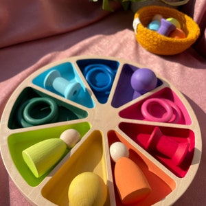 Può includere: Un vassoio di legno color arcobaleno con varie forme e colori. Il vassoio ha 12 scomparti, ognuno con un colore e una forma diversi. Le forme includono un cerchio, un quadrato, un triangolo, una stella, un cuore e una mezzaluna. I colori includono rosso, arancione, giallo, verde, blu, viola, rosa, marrone, nero, bianco, grigio e beige.