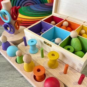 DiDibox, caja de madera con muñeca, bolillos, anillos y pelota. Juego Montessori, juego de clasificación.