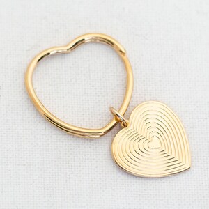 May include: A gold-colored keychain with a heart-shaped design. The keychain has a circular ring with a heart-shaped pendant. The pendant features concentric heart shapes, creating a layered effect. The keychain is on a white surface.