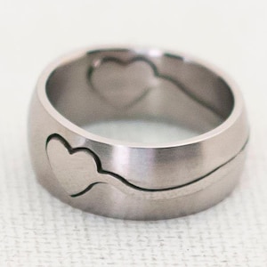 Puede incluir: Un anillo de rompecabezas de plata con dos corazones entrelazados. El anillo está hecho de metal y tiene un acabado liso y pulido.