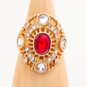 Könnte beinhalten: Ein goldener Ring mit einem großen roten Edelstein, umgeben von kleineren klaren Edelsteinen.