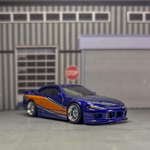 May include: A die-cast model sports car in a deep metallic blue finish with a prominent orange and white side stripe graphic. The vehicle features silver multi-spoke wheels, a low-profile stance, and clear windows, capturing the design of a performance coupe.