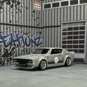 May include: A silver diecast model car with black racing stripes and a white circle on the side. The car is parked in a garage setting with graffiti art on the wall. The text "Liberty Walk" is visible.