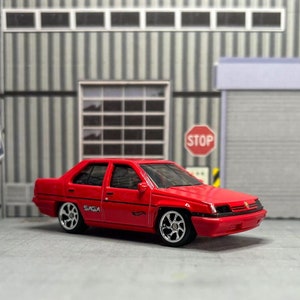 May include: A red toy car model of a Proton Saga, a Malaysian car, with black trim and silver wheels. The car is parked in front of a garage door with a "STOP" sign.