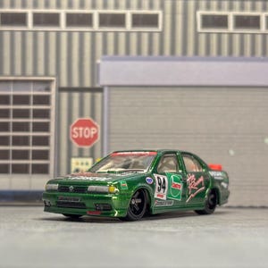May include: A green and black toy car with racing decals and the number 94 on the side. The car is a sedan style and has black wheels. The background includes a garage door and a stop sign.